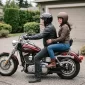 A rider and passenger seated on a parked cruiser in a driveway, both in helmets, the passenger's feet on the floorboards
